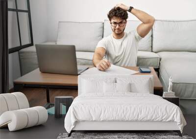 Hispanic man doing papers at home confuse and wondering about question. uncertain with doubt, thinking with hand on head. pensive concept. Wall mural