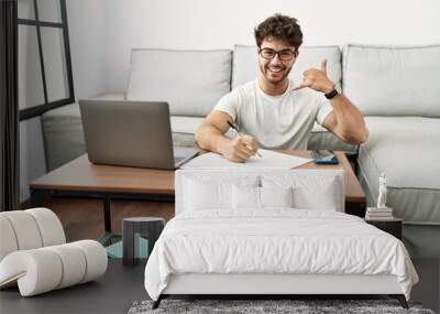 Hispanic man doing papers at home smiling doing phone gesture with hand and fingers like talking on the telephone. communicating concepts. Wall mural