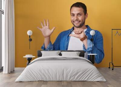 Hispanic man standing over yellow background showing and pointing up with fingers number six while smiling confident and happy. Wall mural