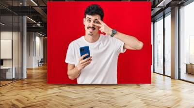 Hispanic man using smartphone over red background doing peace symbol with fingers over face, smiling cheerful showing victory Wall mural