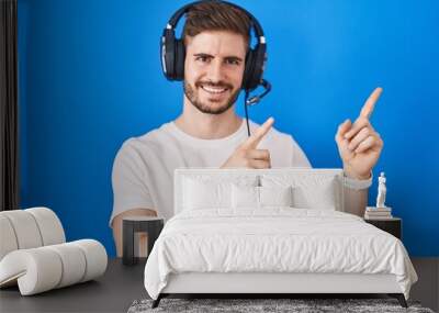 Hispanic man with beard listening to music wearing headphones smiling and looking at the camera pointing with two hands and fingers to the side. Wall mural