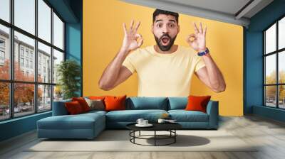 Hispanic man with beard standing over yellow background looking surprised and shocked doing ok approval symbol with fingers. crazy expression Wall mural