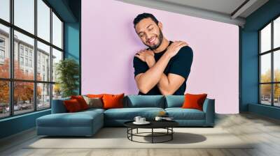 Hispanic man with beard wearing casual black t shirt hugging oneself happy and positive, smiling confident. self love and self care Wall mural