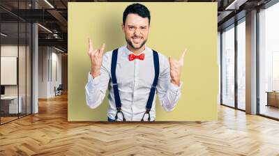 Hispanic man with beard wearing hipster look with bow tie and suspenders shouting with crazy expression doing rock symbol with hands up. music star. heavy concept. Wall mural