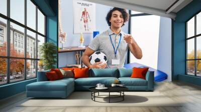 Hispanic man with curly hair working as football physiotherapist pointing to you and the camera with fingers, smiling positive and cheerful Wall mural