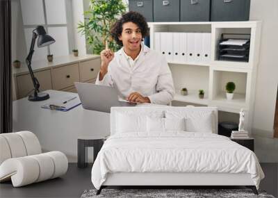 Hispanic man with curly hair working at optician office smiling with an idea or question pointing finger up with happy face, number one Wall mural