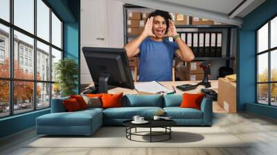 Hispanic man with curly hair working at small business ecommerce smiling cheerful playing peek a boo with hands showing face. surprised and exited Wall mural