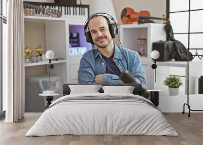 Hispanic man with headphones smiling in a music studio, surrounded by instruments and recording equipment. Wall mural