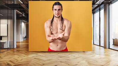 Hispanic man with long hair standing shirtless over yellow background smiling with hands palms together receiving or giving gesture. hold and protection Wall mural