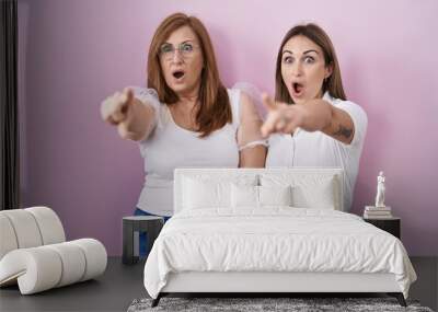 Hispanic mother and daughter wearing casual white t shirt over pink background pointing with finger surprised ahead, open mouth amazed expression, something on the front Wall mural