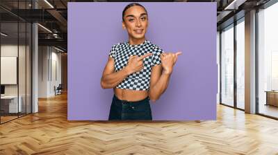 Hispanic transgender man wearing make up and long hair wearing modern clothes smiling and looking at the camera pointing with two hands and fingers to the side. Wall mural