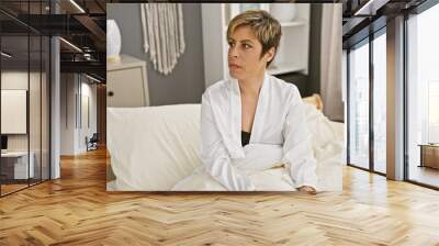 Hispanic woman with short blonde hair wearing a white robe sits contemplatively on a bed indoors. Wall mural