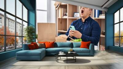 Hispanic young man eating take away food using smartphone angry and mad screaming frustrated and furious, shouting with anger looking up. Wall mural