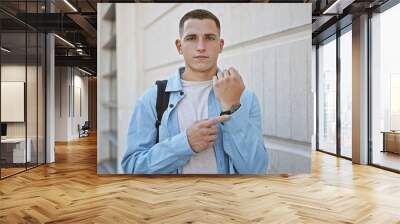 Hispanic young man in casual attire pointing at his watch on a city street, looking punctual and ready. Wall mural