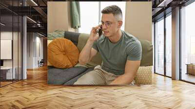 Hispanic young man in casual attire sitting on a sofa, talking on smartphone in a modern home interior. Wall mural