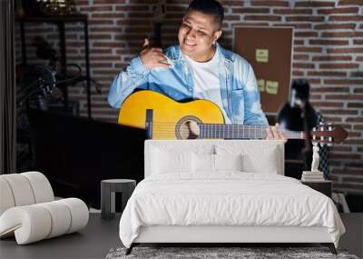 Hispanic young man playing classic guitar at music studio smiling doing phone gesture with hand and fingers like talking on the telephone. communicating concepts. Wall mural