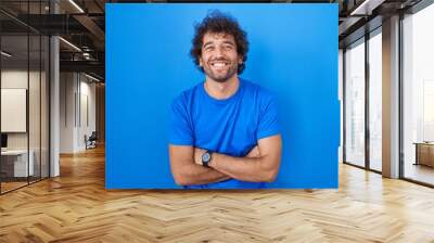 Hispanic young man standing over blue background happy face smiling with crossed arms looking at the camera. positive person. Wall mural