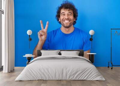 Hispanic young man standing over blue background showing and pointing up with fingers number two while smiling confident and happy. Wall mural