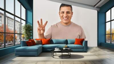 Hispanic young man standing over white background showing and pointing up with fingers number three while smiling confident and happy. Wall mural