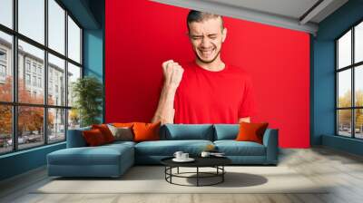 Hispanic young man wearing casual red t shirt celebrating surprised and amazed for success with arms raised and eyes closed Wall mural