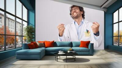 Hispanic young man wearing doctor uniform very happy and excited doing winner gesture with arms raised, smiling and screaming for success. celebration concept. Wall mural