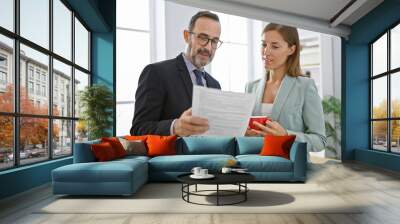 In the heart of a professional morning, two serious business workers, standing together in the office interior, savoring espresso while ardently reading a success-driven work document. Wall mural