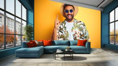 Indian man on vacation wearing floral shirt hat sunglasses over isolated yellow background pointing finger up with successful idea. Exited and happy. Number one. Wall mural