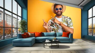 Indian man on vacation wearing floral shirt hat sunglasses over isolated yellow background smiling and looking at the camera pointing with two hands and fingers to the side. Wall mural