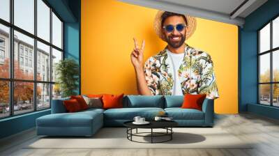 Indian man on vacation wearing floral shirt hat sunglasses over isolated yellow background smiling with happy face winking at the camera doing victory sign. Number two. Wall mural