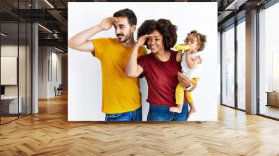 Interracial young family of black mother and hispanic father with daughter very happy and smiling looking far away with hand over head. searching concept. Wall mural