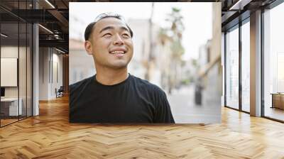 Joyful young chinese man, confidently smiling, looking to the sky while standing in the urban street Wall mural