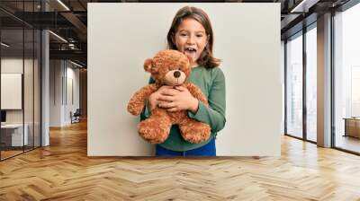 Little beautiful girl hugging teddy bear sticking tongue out happy with funny expression. Wall mural