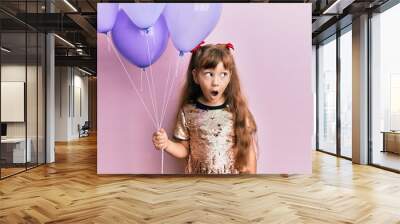 Little caucasian girl kid holding balloons scared and amazed with open mouth for surprise, disbelief face Wall mural