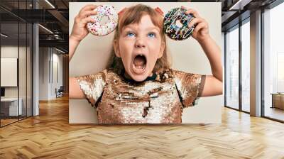 Little caucasian girl kid holding tasty colorful doughnuts angry and mad screaming frustrated and furious, shouting with anger looking up. Wall mural