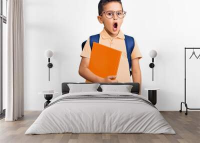 Little cute boy kid wearing school bag and holding book scared and amazed with open mouth for surprise, disbelief face Wall mural