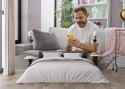 Mature bearded man in casual attire using smartphone on a sofa indoors, embodying leisure at home. Wall mural