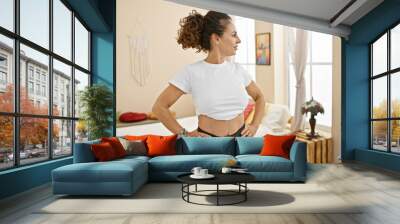 Mature hispanic woman with curly hair standing confidently in a cozy bedroom, hands on hips, wearing casual attire. Wall mural