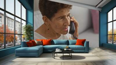 Mature hispanic woman with short hair talking on a landline phone in her living room, showcasing an indoor and home setting. Wall mural