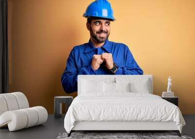 Mechanic man with beard wearing blue uniform and safety helmet over yellow background laughing nervous and excited with hands on chin looking to the side Wall mural