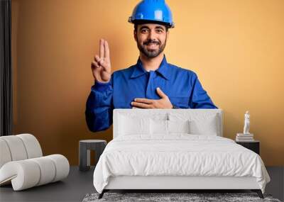 Mechanic man with beard wearing blue uniform and safety helmet over yellow background smiling swearing with hand on chest and fingers up, making a loyalty promise oath Wall mural