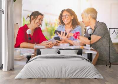 Meeting of middle age women having lunch and drinking coffee. Mature friends smiling happy using smartphone at home on a sunny day Wall mural