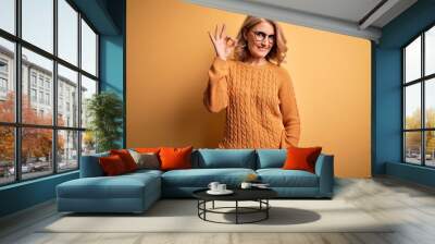 Middle age beautiful blonde woman wearing casual sweater and glasses over yellow background smiling positive doing ok sign with hand and fingers. Successful expression. Wall mural