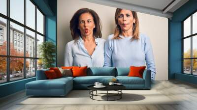 Middle age beautiful couple of sisters standing over isolated white background making fish face with lips, crazy and comical gesture. Funny expression. Wall mural