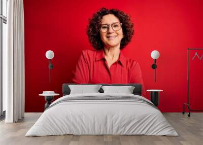 Middle age beautiful curly hair woman wearing casual shirt and glasses over red background happy face smiling with crossed arms looking at the camera. Positive person. Wall mural