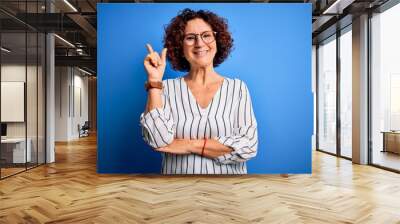 Middle age beautiful curly hair woman wearing casual striped shirt over isolated background with a big smile on face, pointing with hand and finger to the side looking at the camera. Wall mural