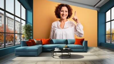 Middle age beautiful curly hair woman wearing casual summer dress over yellow background pointing finger up with successful idea. Exited and happy. Number one. Wall mural