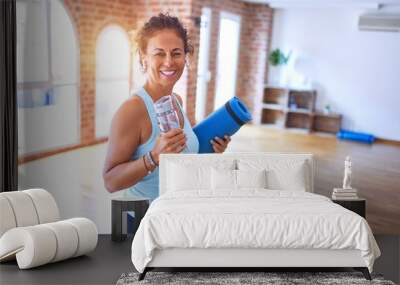 Middle age beautiful sportwoman smiling happy and confident. Standing with smile on face holding mat and bottle of water before doing exercise at gym Wall mural