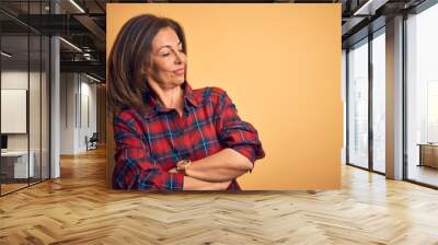 Middle age beautiful woman wearing casual shirt standing over isolated yellow background looking to the side with arms crossed convinced and confident Wall mural