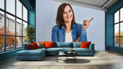 Middle age brunette woman wearing casual denim jacket over isolated background with a big smile on face, pointing with hand and finger to the side looking at the camera. Wall mural