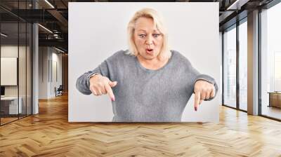 Middle age caucasian woman standing over white background pointing down with fingers showing advertisement, surprised face and open mouth Wall mural